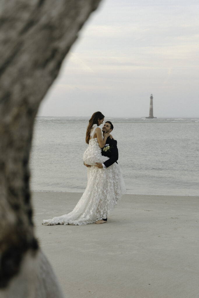 A couple on a beach in South Carolina, one of the best places to elope on the east coast. They're in their elopement attire, and the groom is holding the bride, with a lighthouse in the background.