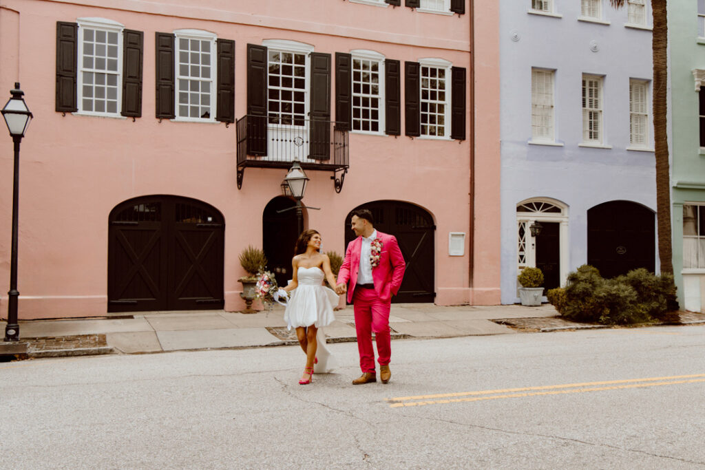 A couple standing in front of Rainbow Row in Charleston, one of the best places to elope on the east coast for urban vibes! They're in their elopement attire, holding hands and looking at each other as they walk across the street.